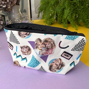 May include: White canvas zippered pouch with an 80s theme. The pouch features a repeating pattern of geometric shapes, cassette tapes, and images of people. The colours in the pattern are blue, purple, black, and white.