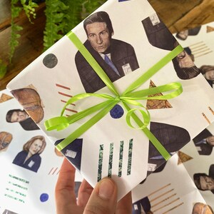 May include: White gift wrap with a green ribbon. The wrapping paper features a repeating pattern of illustrations of characters from the TV show "Twin Peaks" with geometric shapes in red, blue, and brown.