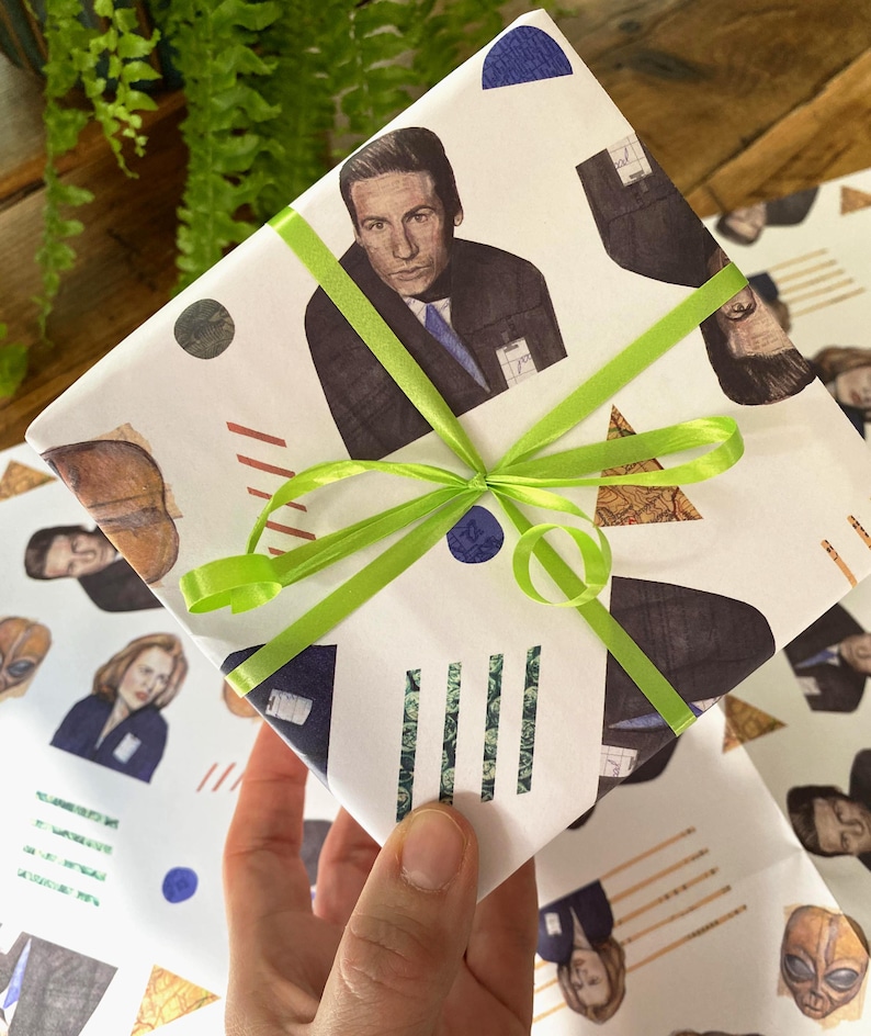 May include: White gift wrap with a green ribbon. The wrapping paper features a repeating pattern of illustrations of characters from the TV show "Twin Peaks" with geometric shapes in red, blue, and brown.