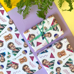 May include: White wrapping paper with a repeating pattern of a man's portrait, a dog, a garden fork, and geometric shapes. A wrapped gift is tied with a green ribbon. The background includes yellow, purple, and green foliage.