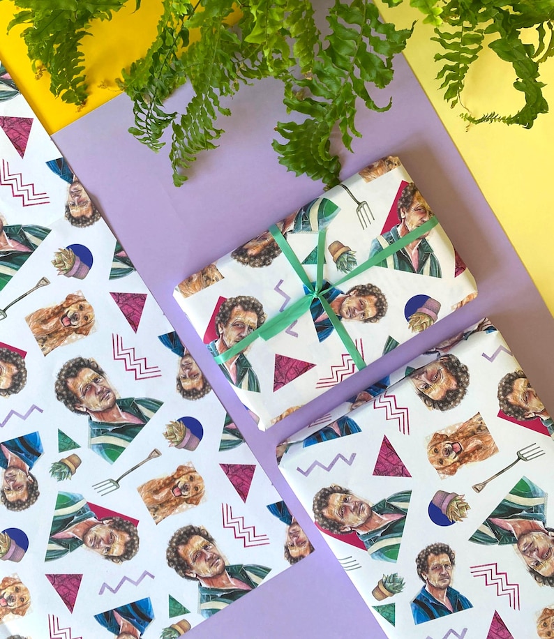 May include: White wrapping paper with a repeating pattern of a man's portrait, a dog, a garden fork, and geometric shapes. A wrapped gift is tied with a green ribbon. The background includes yellow, purple, and green foliage.