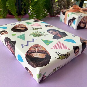 May include: A wrapped gift with white paper decorated with repeating illustrations of faces, geometric shapes, and a fern. The gift is on a purple surface. Another gift box is visible in the background.