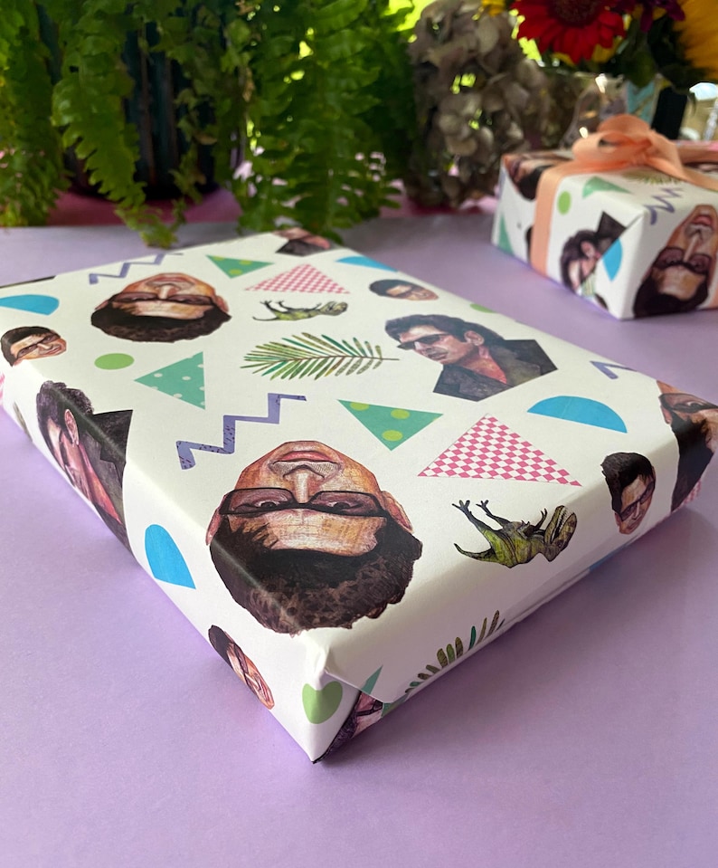 May include: A wrapped gift with white paper decorated with repeating illustrations of faces, geometric shapes, and a fern. The gift is on a purple surface. Another gift box is visible in the background.