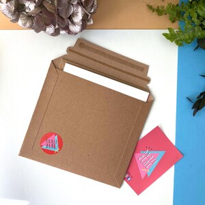 May include: A brown paper envelope, partially open to show white paper inside. A pink card with the text "ANNA DEAL" and a geometric design is next to the envelope. A red sticker with the same text and design is on the envelope.