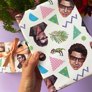 May include: White wrapping paper with a repeating pattern of a man's face, dinosaurs, geometric shapes, and palm leaves. A second wrapped gift with a peach ribbon is visible. The background includes flowers and greenery.