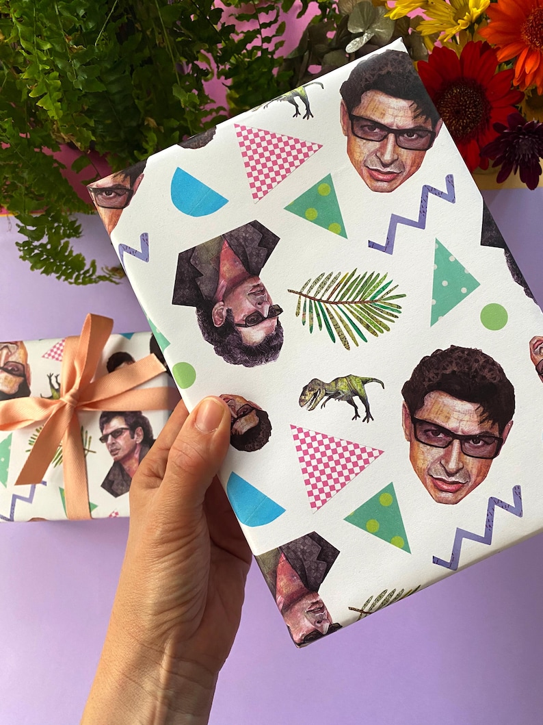 May include: White wrapping paper with a repeating pattern of a man's face, dinosaurs, geometric shapes, and palm leaves. A second wrapped gift with a peach ribbon is visible. The background includes flowers and greenery.
