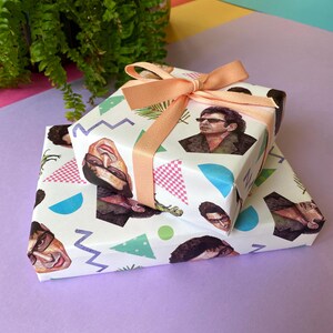 May include: Two wrapped gifts with white paper featuring a repeating pattern of a person's face, geometric shapes, and palm fronds. The gifts are tied with a peach-coloured ribbon. The gifts are stacked on a purple surface.