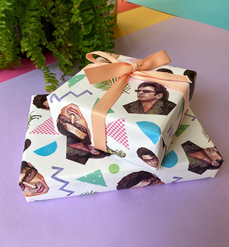 May include: Two wrapped gifts with white paper featuring a repeating pattern of a person's face, geometric shapes, and palm fronds. The gifts are tied with a peach-coloured ribbon. The gifts are stacked on a purple surface.