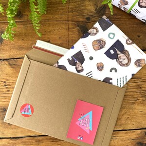 May include: A brown paper envelope with a white and blue patterned wrapping paper inside. The wrapping paper features a repeating pattern of faces and geometric shapes. The envelope has a red and white sticker with the text "an all deal" on it.