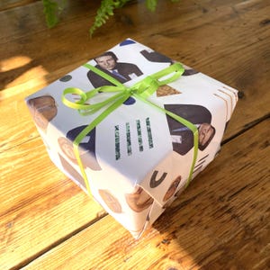 May include: A white gift box wrapped in paper with a collage of images and text. The box is tied with a green ribbon.