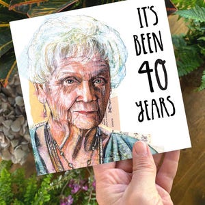 May include: A square greeting card featuring a watercolour portrait of an elderly woman with white hair. The card reads "IT'S BEEN 40 YEARS" in black handwritten font. The card is held in a hand.