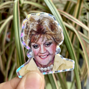 May include: A sticker of a woman with blonde hair wearing a yellow shirt and a pearl necklace. The sticker has a holographic finish.