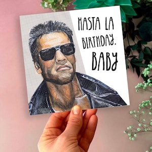 May include: A birthday card featuring a portrait of a man wearing sunglasses. The card has the text "Hasta La Birthday, Baby" in black lettering. The background is a light pink color with green plants.