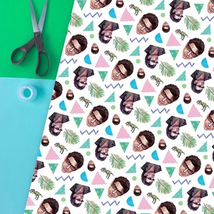 May include: White wrapping paper featuring a repeating pattern of a man's face, dinosaurs, and geometric shapes in blue, pink, and green. Scissors and tape are on a green and blue background.