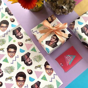 May include: A wrapped gift with peach ribbon, featuring white wrapping paper printed with a repeating pattern of a man's face, dinosaurs, and geometric shapes. Matching wrapping paper and a pink card are also present.