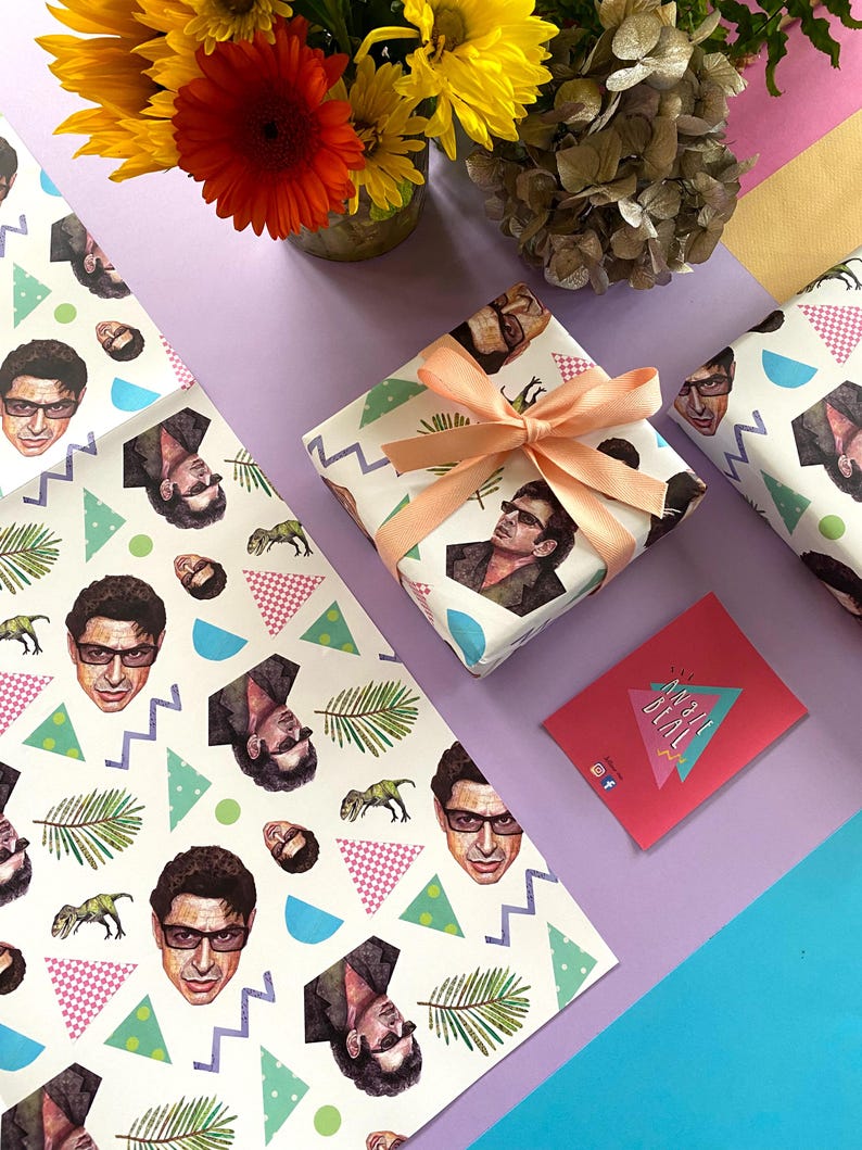 May include: A wrapped gift with peach ribbon, featuring white wrapping paper printed with a repeating pattern of a man's face, dinosaurs, and geometric shapes. Matching wrapping paper and a pink card are also present.