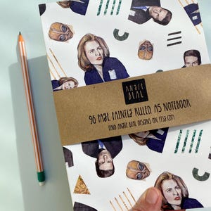 May include: A white A5 notebook with illustrations of characters and alien masks. The notebook has a brown band with the text "96 PAGE FAINTLY RULED. A5 NOTEBOOK" and the brand name "ANGIE DEAL". A pencil with orange and white stripes is next to the notebook.