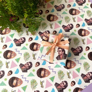 May include: Wrapping paper featuring a repeating pattern of a man's face, dinosaurs, and geometric shapes. A wrapped gift with a peach ribbon and bow is in the center. Ferns and flowers are in the background.
