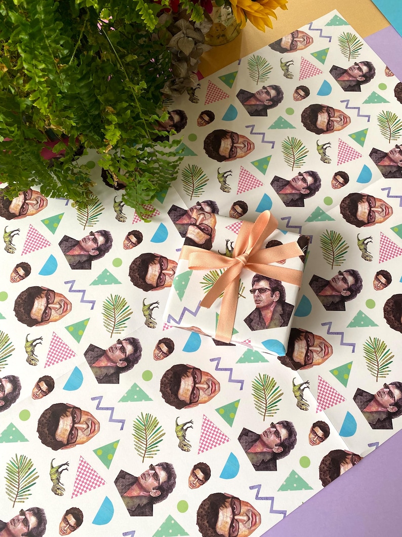 May include: Wrapping paper featuring a repeating pattern of a man's face, dinosaurs, and geometric shapes. A wrapped gift with a peach ribbon and bow is in the center. Ferns and flowers are in the background.