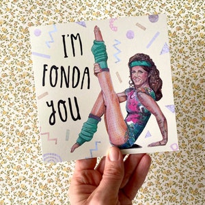 May include: A greeting card with a retro 80s aesthetic featuring a woman in a pink and green leotard and leg warmers. The card says "I'm Fonda You".