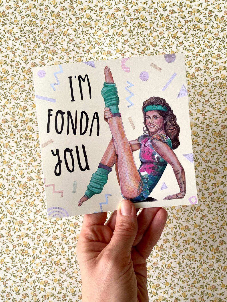 May include: A greeting card with a retro 80s aesthetic featuring a woman in a pink and green leotard and leg warmers. The card says "I'm Fonda You".