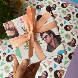 May include: A wrapped present with patterned paper featuring illustrations of a person wearing sunglasses. The gift is tied with a peach-coloured ribbon. The background includes additional patterned paper and a bouquet of flowers.