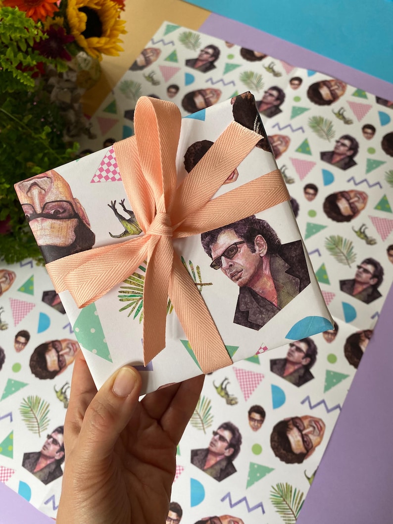May include: A wrapped present with patterned paper featuring illustrations of a person wearing sunglasses. The gift is tied with a peach-coloured ribbon. The background includes additional patterned paper and a bouquet of flowers.