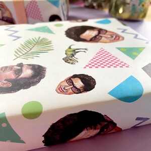 May include: Gift wrap with a repeating pattern of a person's face, a dinosaur, and geometric shapes in blue, green, pink, and grey. Another gift box with a pink bow is in the background.