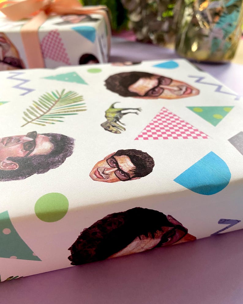 May include: Gift wrap with a repeating pattern of a person's face, a dinosaur, and geometric shapes in blue, green, pink, and grey. Another gift box with a pink bow is in the background.