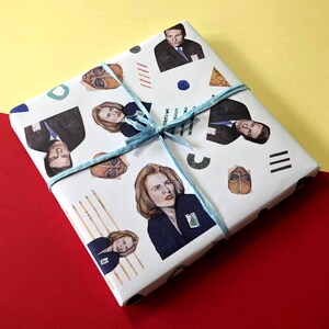 May include: White gift wrap with a blue ribbon and a repeating pattern of characters from the TV show The X-Files. The characters include Mulder, Scully, and the Smoking Man.