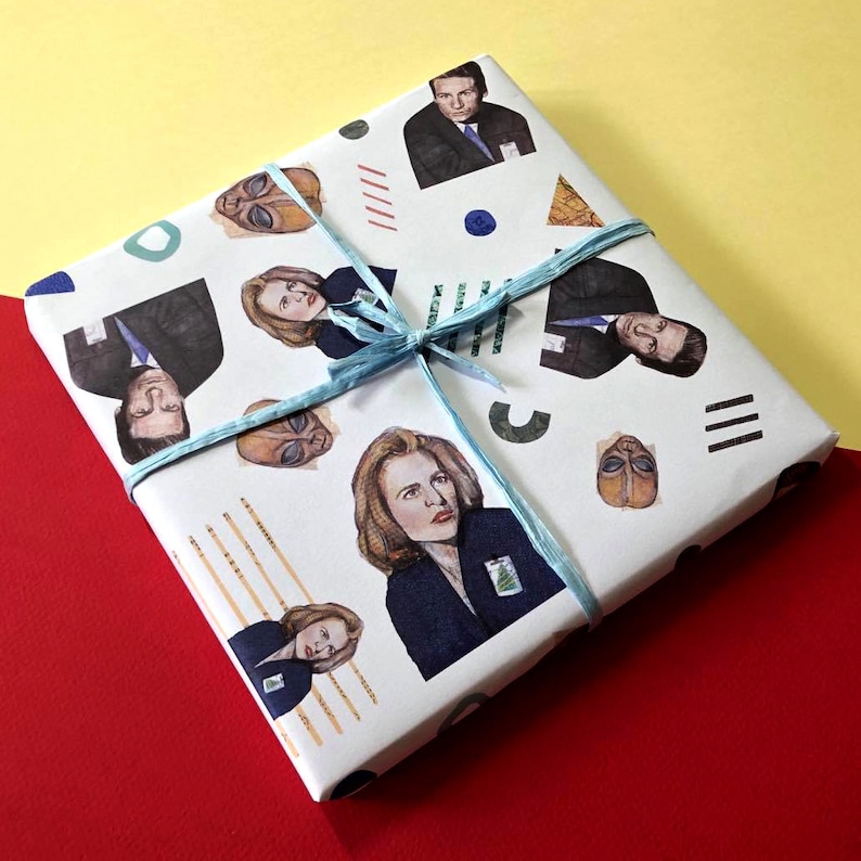 May include: White gift wrap with a blue ribbon and a repeating pattern of characters from the TV show The X-Files. The characters include Mulder, Scully, and the Smoking Man.