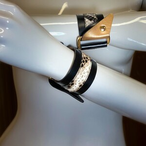 May include: Two black leather bracelets with gold accents and a snakeskin pattern. The bracelets are worn on a mannequin's arms.