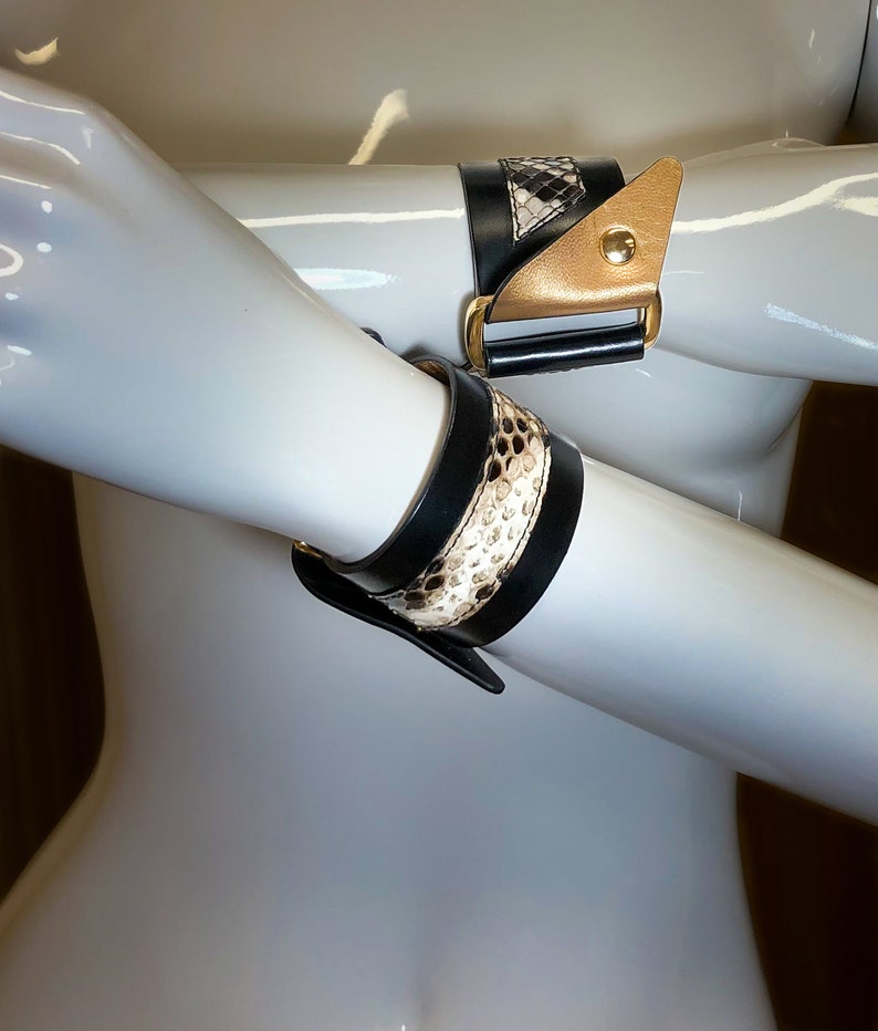 May include: Two black leather bracelets with gold accents and a snakeskin pattern. The bracelets are worn on a mannequin's arms.