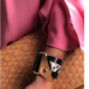 May include: A black leather bracelet with a gold buckle and a snakeskin pattern. The bracelet is worn on a person's wrist.