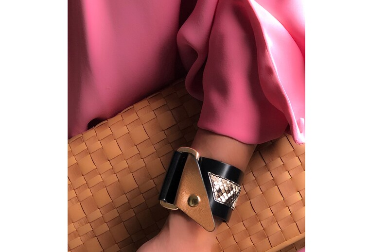 May include: A black leather bracelet with a gold buckle and a snakeskin pattern. The bracelet is worn on a person's wrist.