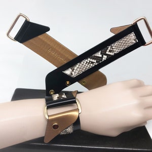 May include: A black and gold leather bracelet with a snakeskin pattern. The bracelet has a buckle closure and is adjustable.