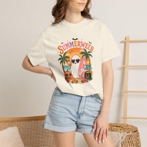 Może przedstawiać: Kremowy t-shirt z krótkim rękawem z nadrukiem graficznym z napisem "SUMMERWEEN" nad duchem w okularach przeciwsłonecznych w kształcie serca, trzymającym rożek lodowy i deskę surfingową, w otoczeniu palm, dyń i kwiatów.