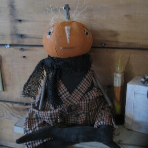 K&ouml;nnte beinhalten: Eine handgemachte K&uuml;rbispuppe mit einem schwarzen und braunen karierten Outfit und einer schwarzen Wollper&uuml;cke. Die Puppe sitzt auf einem Holzregal mit einer Kerze und einer Kiste dahinter.