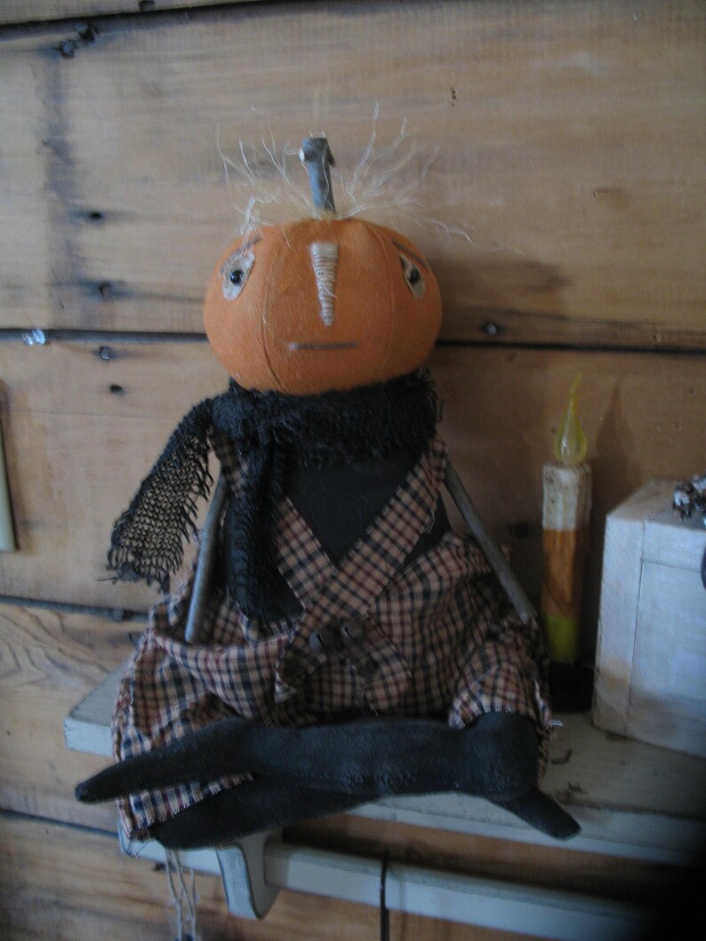 K&ouml;nnte beinhalten: Eine handgemachte K&uuml;rbispuppe mit einem schwarzen und braunen karierten Outfit und einer schwarzen Wollper&uuml;cke. Die Puppe sitzt auf einem Holzregal mit einer Kerze und einer Kiste dahinter.