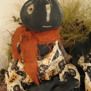 K&ouml;nnte beinhalten: Eine handgemachte Stoffpuppe mit einem schwarzen Stoffkopf und einem braunen Sackleinen-Schal. Die Puppe tr&auml;gt ein wei&szlig;es Stoffkleid mit einem schwarzen und orangefarbenen K&uuml;rbis-Muster. Die Puppe sitzt auf einer braunen Oberfl&auml;che.