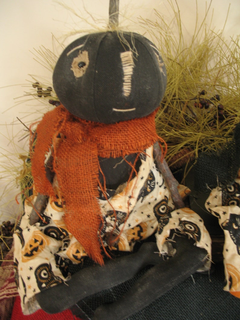 K&ouml;nnte beinhalten: Eine handgemachte Stoffpuppe mit einem schwarzen Stoffkopf und einem braunen Sackleinen-Schal. Die Puppe tr&auml;gt ein wei&szlig;es Stoffkleid mit einem schwarzen und orangefarbenen K&uuml;rbis-Muster. Die Puppe sitzt auf einer braunen Oberfl&auml;che.