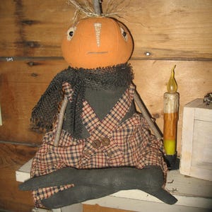 K&ouml;nnte beinhalten: Eine handgemachte K&uuml;rbispuppe, die eine schwarze und rote karierter Latzhose und einen schwarzen Schal tr&auml;gt. Die Puppe sitzt auf einem Holzregal mit einer Kerze daneben.