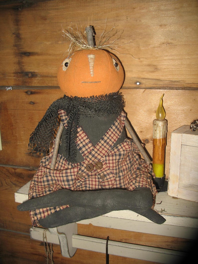 K&ouml;nnte beinhalten: Eine handgemachte K&uuml;rbispuppe, die eine schwarze und rote karierter Latzhose und einen schwarzen Schal tr&auml;gt. Die Puppe sitzt auf einem Holzregal mit einer Kerze daneben.