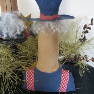 May include: A handmade rag doll with a blue hat, red and white checkered overalls, and a white beard. The doll is sitting on a bed of pine needles.