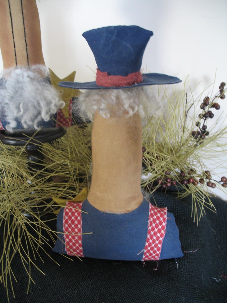 May include: A handmade rag doll with a blue hat, red and white checkered overalls, and a white beard. The doll is sitting on a bed of pine needles.