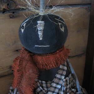 K&ouml;nnte beinhalten: Eine schwarze Stoffpuppe mit einem karierten Kleid und einem braunen Sackleinen-Schal. Die Puppe hat ein einfaches Gesicht mit gestickten Augen und einem Mund. Die Puppe hat stroh&auml;hnliches Haar auf dem Kopf.