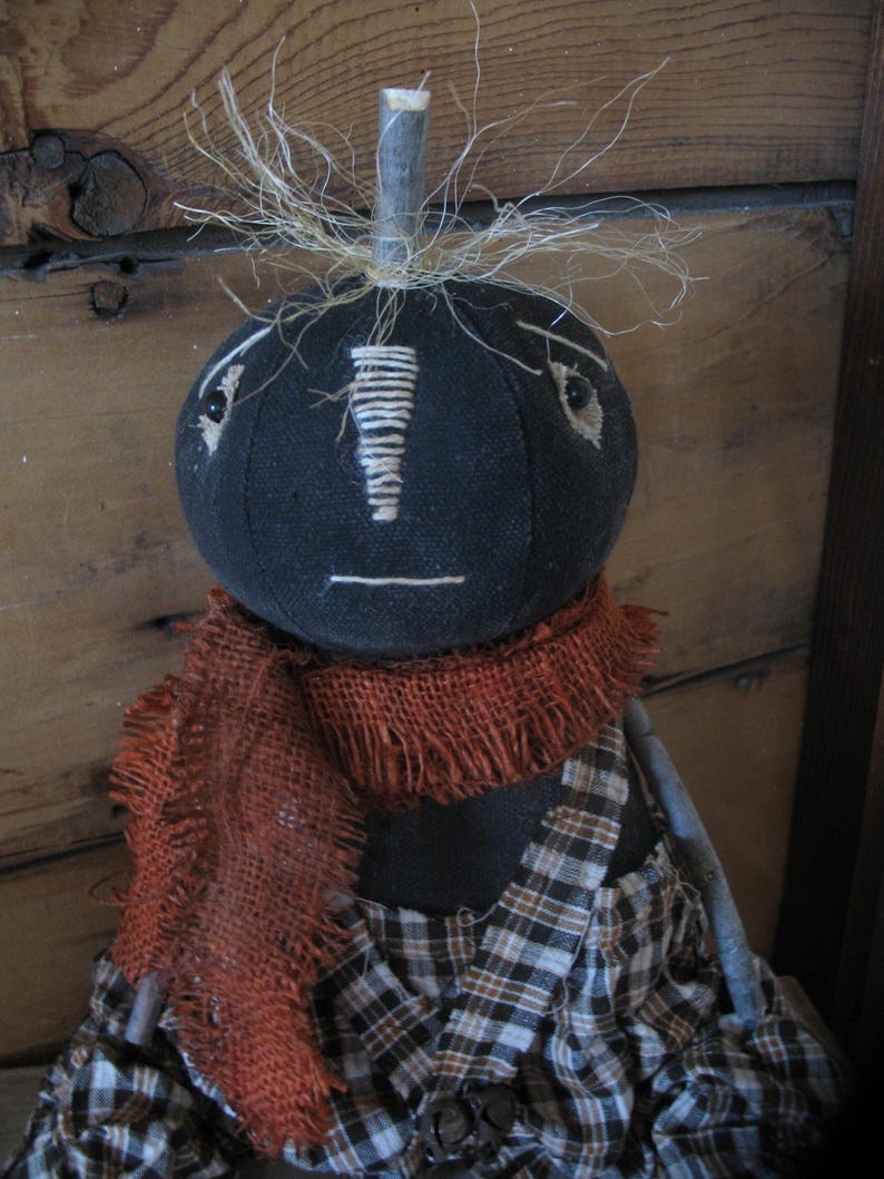 K&ouml;nnte beinhalten: Eine schwarze Stoffpuppe mit einem karierten Kleid und einem braunen Sackleinen-Schal. Die Puppe hat ein einfaches Gesicht mit gestickten Augen und einem Mund. Die Puppe hat stroh&auml;hnliches Haar auf dem Kopf.