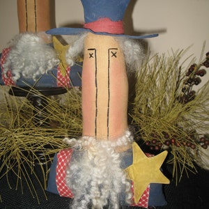 May include: Two handmade rag dolls dressed in patriotic red, white, and blue clothing. The dolls have white yarn beards and are wearing hats. The doll on the right is wearing a blue top hat with a red band. The doll on the left is wearing a blue and white striped hat. Both dolls are standing on a brown surface.