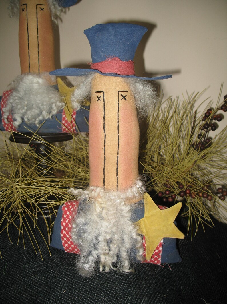 May include: Two handmade rag dolls dressed in patriotic red, white, and blue clothing. The dolls have white yarn beards and are wearing hats. The doll on the right is wearing a blue top hat with a red band. The doll on the left is wearing a blue and white striped hat. Both dolls are standing on a brown surface.