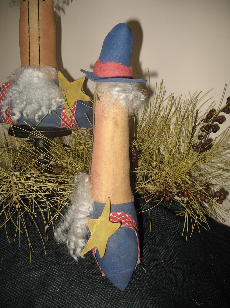 May include: Two handmade rag dolls dressed in red, white, and blue. The doll on the left is wearing a blue hat and the doll on the right is wearing a blue hat with a red band. Both dolls have white yarn hair and are holding a yellow star.